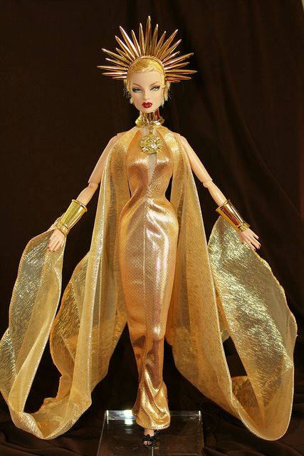 a gold colored doll with an elaborate headpiece and golden dress on display in front of a black backdrop