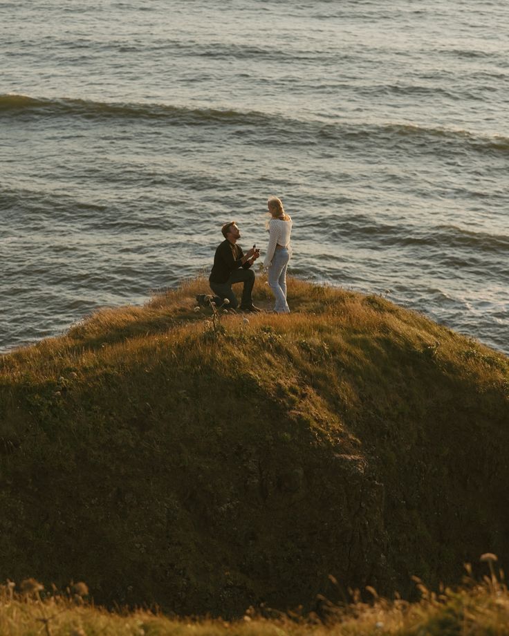 two people sitting on top of a hill next to the ocean