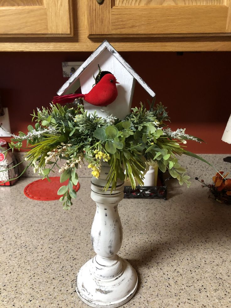 a red bird sitting on top of a vase filled with flowers and greenery next to a kitchen counter