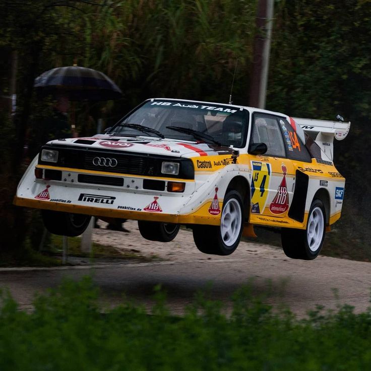 a yellow and white race car is in the air over a dirt road with trees behind it