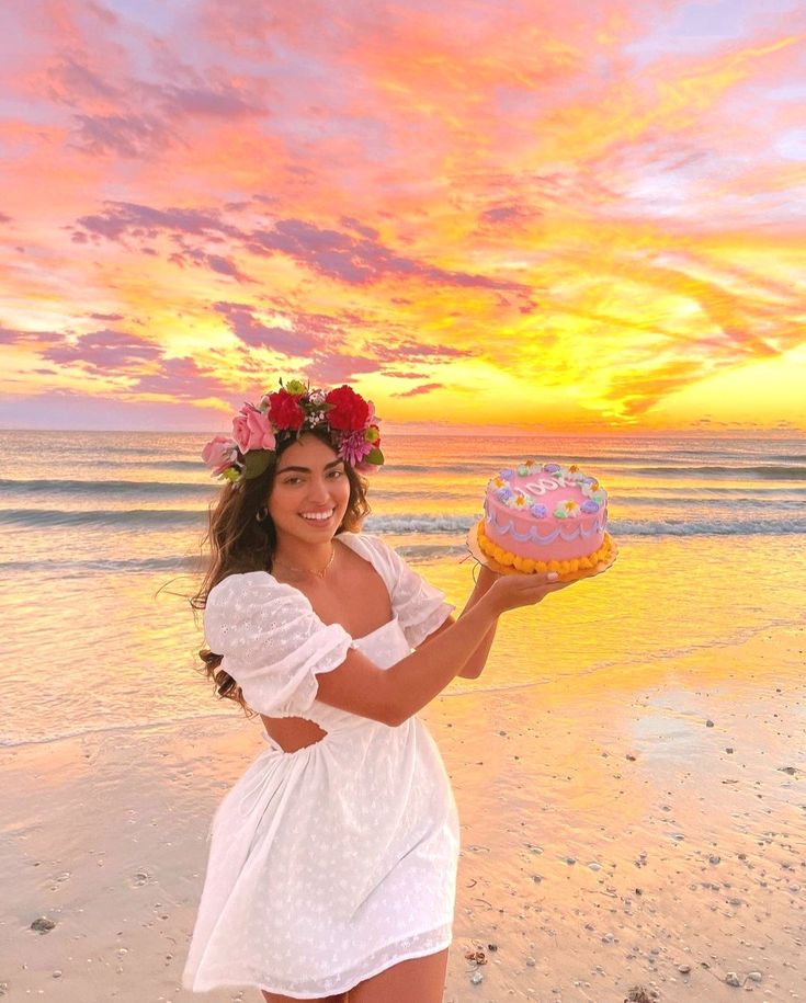 a woman in white dress holding a cake on beach with pink sky and clouds behind her