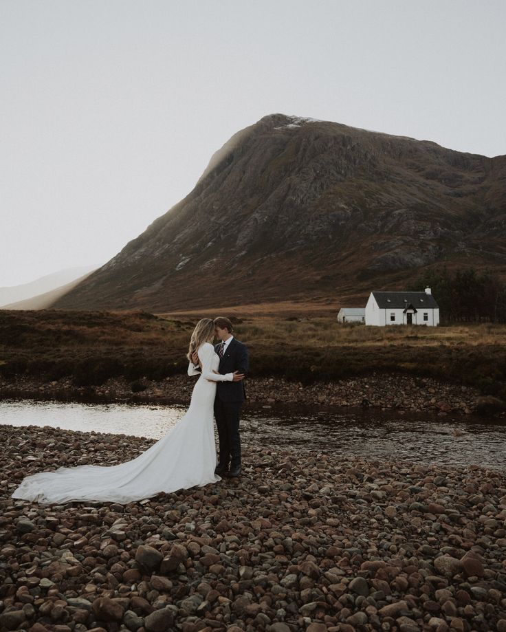 a bride and groom standing on rocks in front of a house with a mountain behind them