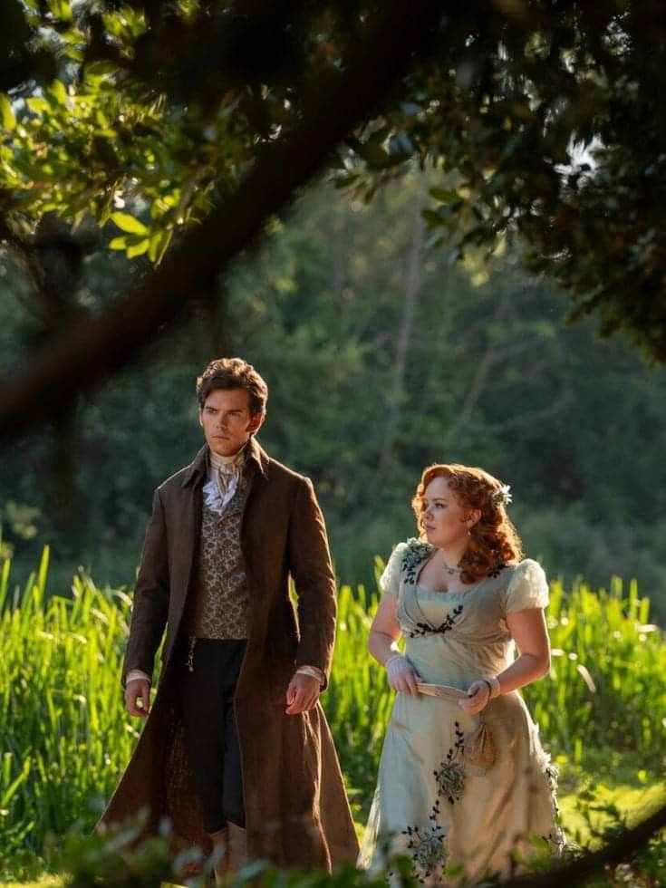 a man and woman dressed in period clothing walking through tall grass with trees behind them