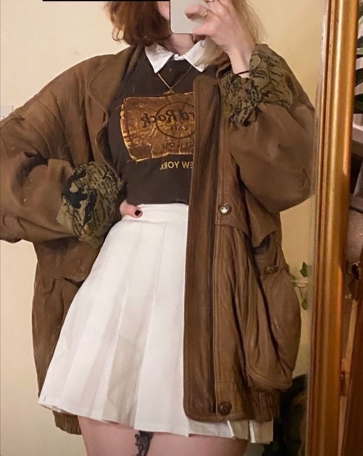 a woman taking a selfie in front of a mirror wearing a brown jacket and white skirt