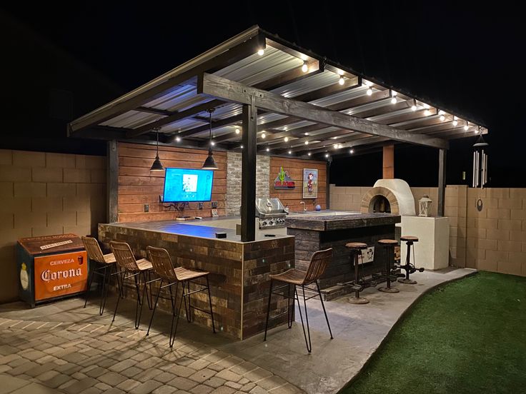 an outdoor kitchen and grill area at night with lights on the roof, bar stools and television