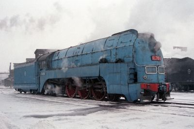 an old blue train traveling down tracks next to snow covered ground with steam pouring out of it