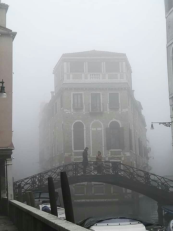 two people standing on a bridge in the middle of a foggy city with buildings