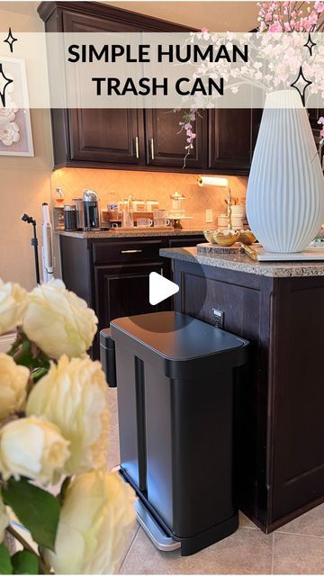 a simple human trash can sits in the middle of a kitchen with flowers on the counter