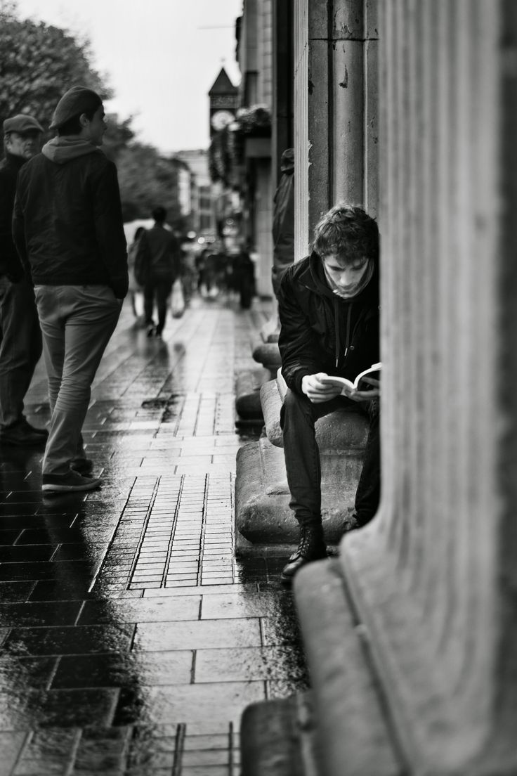 a man sitting on a bench reading a book in the rain while others walk by