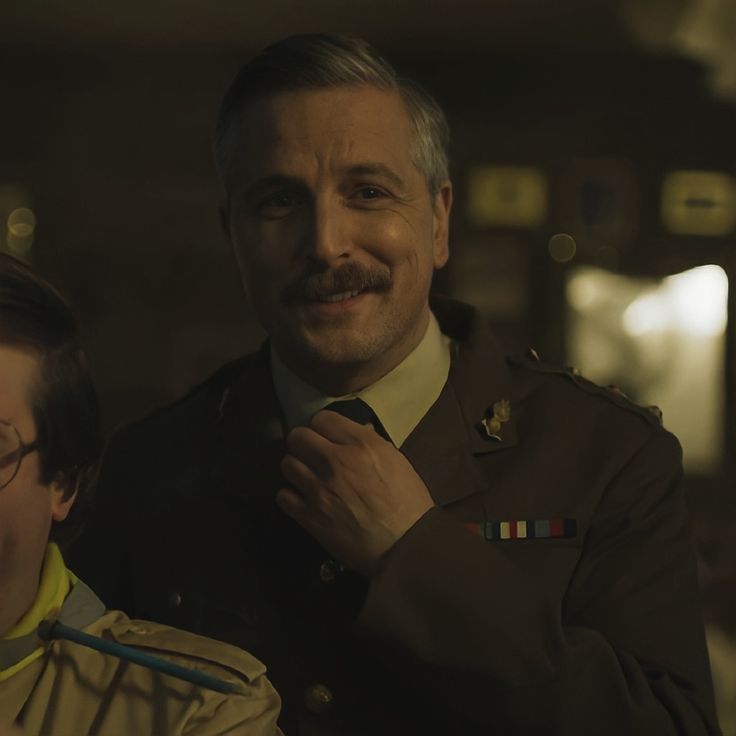 a man adjusting his tie next to another man in uniform and smiling at the camera