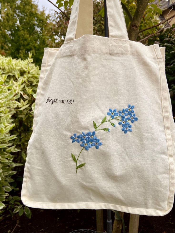 a white tote bag with blue flowers embroidered on the front and writing'forget me not '