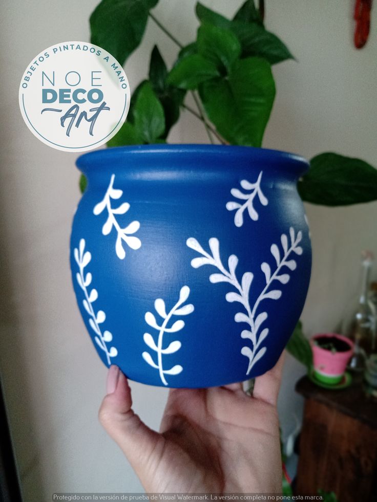 a hand holding a blue pot with white designs on it and a plant in the background