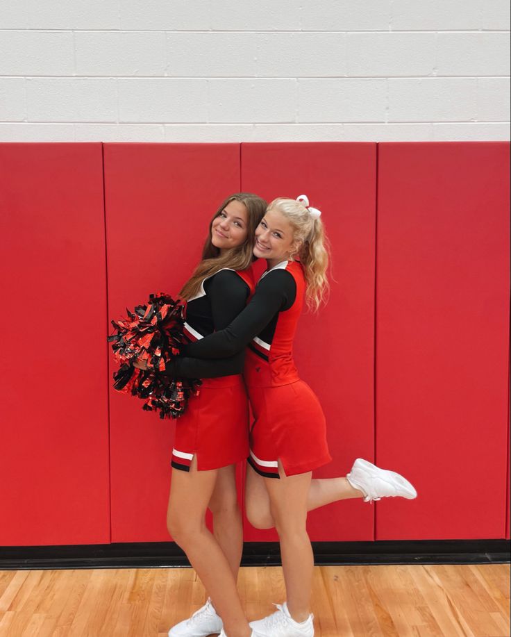 two cheerleaders hugging each other in front of a red wall