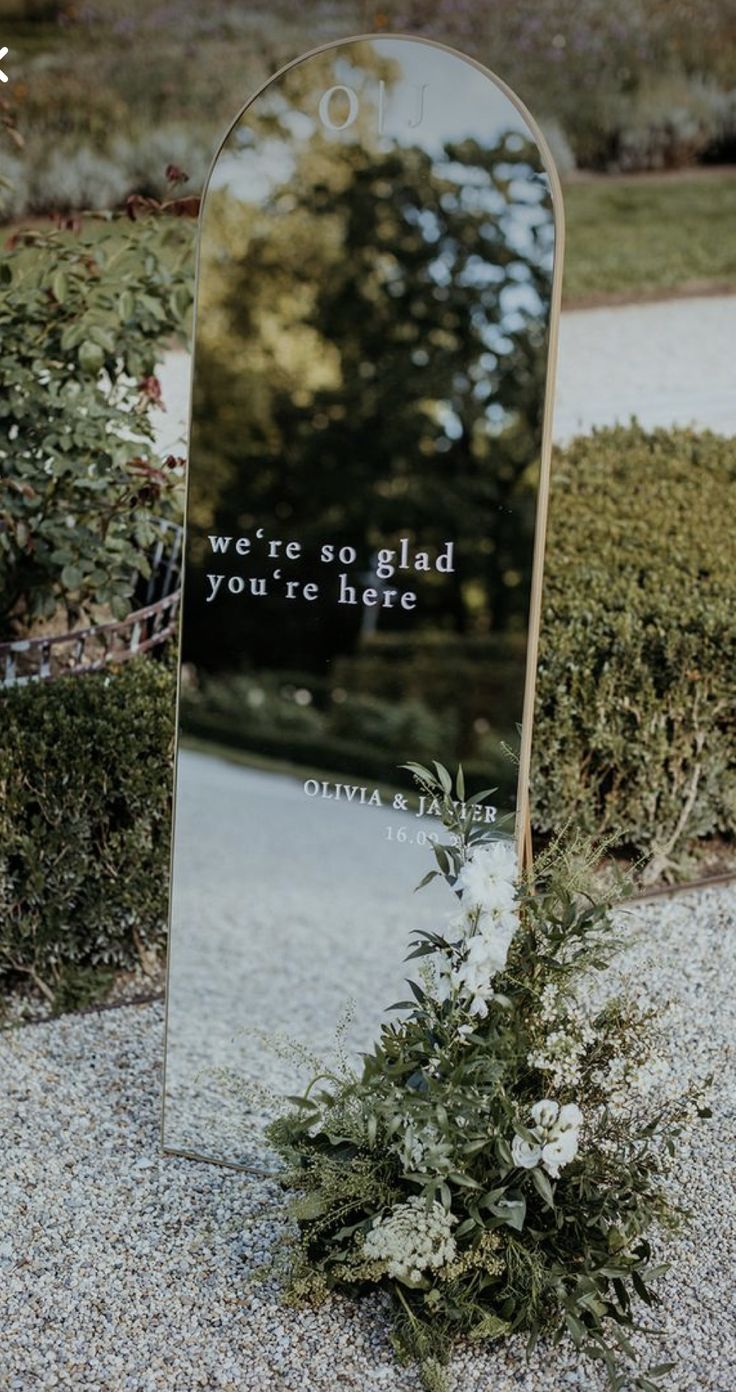 a mirror sitting on the side of a road next to bushes and flowers in front of it