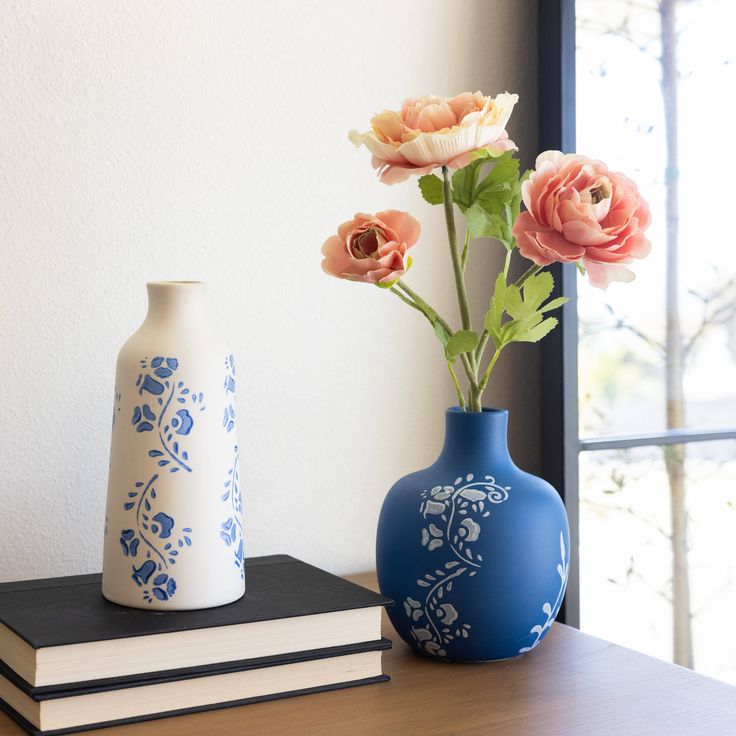 two vases with flowers in them sitting on a table next to books and a window