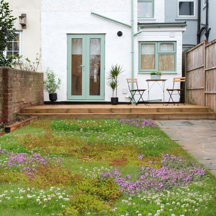 a garden with flowers and grass in front of a white brick house, next to a wooden fence