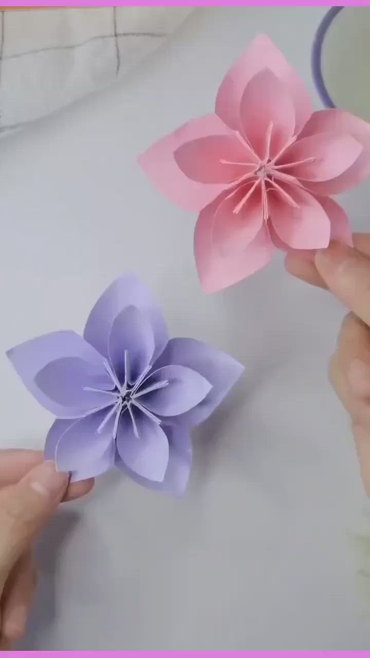 This may contain: two paper flowers being held by someone's hands on a white surface with pink and purple colors