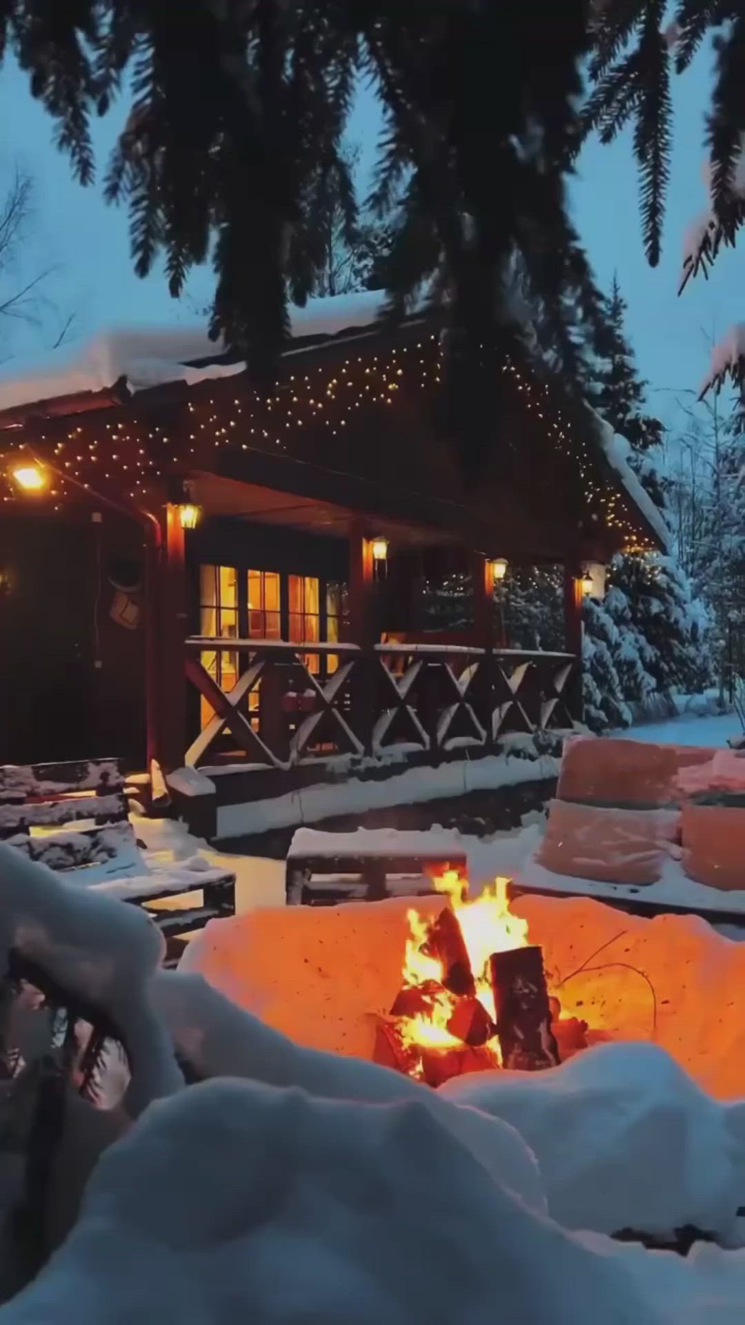 This may contain: a fire pit in the middle of a snowy yard with lights on it and snow covered trees