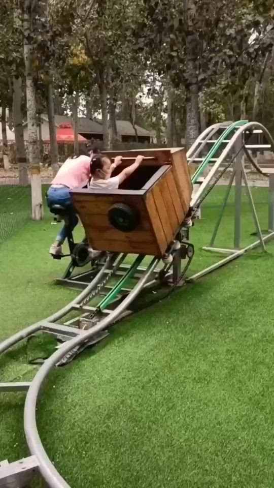 This may contain: two people on a wooden roller coaster in a park with green grass and trees behind them