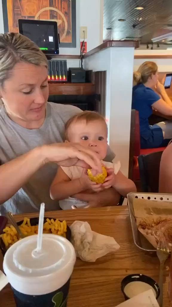 This may contain: a woman holding a baby in her lap while eating corn on the cob at a restaurant