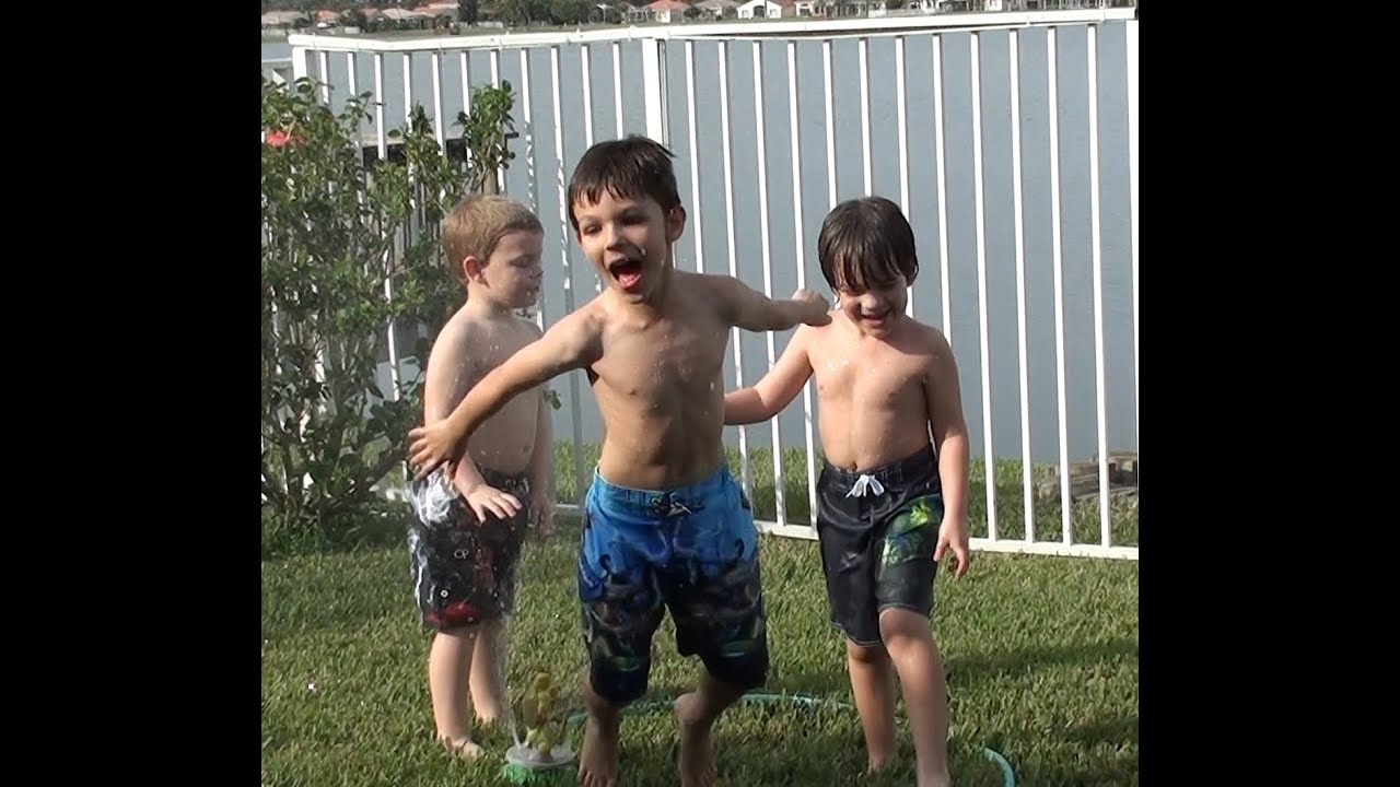 Three Boys Playing In The Backyard Sprinkler With A Hose On A