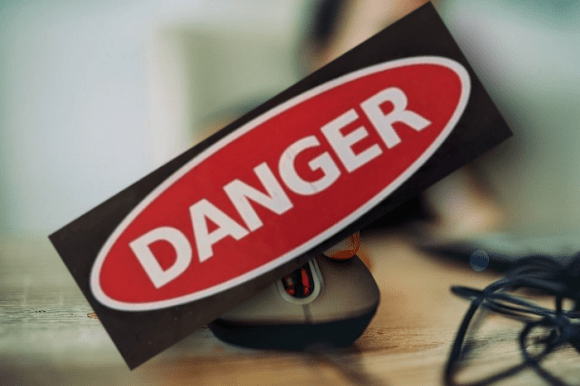 A hand on a multibutton mouse, the body behind it is blurred and out-of-focus; a larger 'DANGER' label in red, white and black, has been superimposed over it. Nenad Stojkovic, CC BY 2.0, modified.