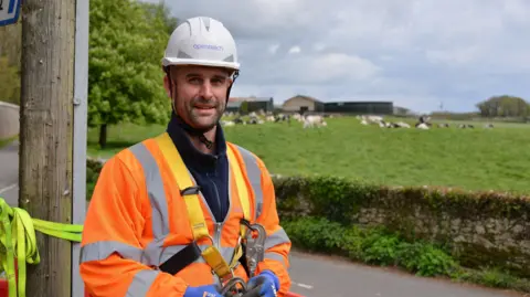 Openreach engineer in a harness, with helmet and high-vis, standing in a rural area