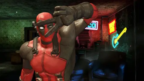 A screenshot of Deadpool from the 2013 computer game showing the peace sign with his fingers.  He is a superhero dressed in a red suit with black over the eyes and gloves.