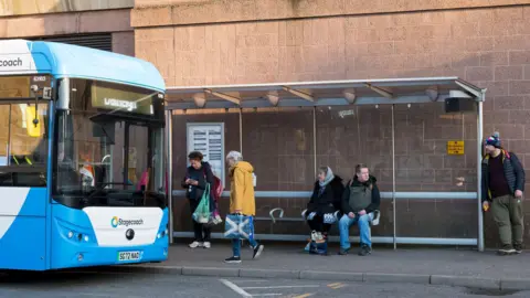 A Stagecoach bus at a bus stop in Inverness. There people sitting and standing waiting for a bus.