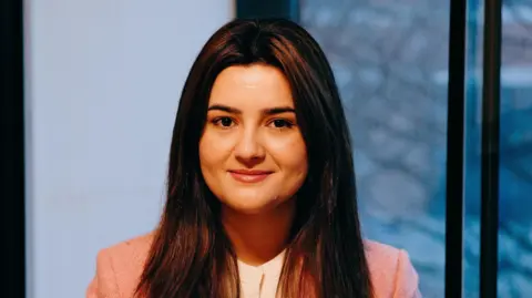 A picture of 22-year-old Mah from Afghanistan who fled to Cardiff when the Taliban took over. She is wearing a pink blazer, standing in front of a glass window.