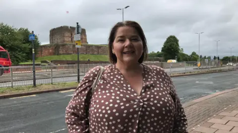 Julie Minns outside the BBC Radio Cumbria office in Carlisle wearing a spotted brown blouse and smiling. There is a road and the castle in the background. 