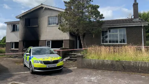 A marked police car parked outside a home which has fire damage on the front wall downstairs and upstairs. The house is a large detached property with brown bricks. There is police tape across the driveway.