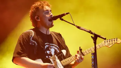 Sam Fender performing on stage. He plays a black and white striped electric guitar while singing into a microphone. He wears a black T-shirt and has short curly fair hair. He's lit by a yellow spotlight.