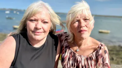 A head and shoulders shot of Lisa Morris and Melanie Leahy standing outdoors on a sunny day looking at the camera. There is water and small boats behind them. Lisa has long hair and is wearing a black top. Melanie also has long hair and is wearing a leopard-skin top. 