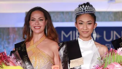 Two women wearing beauty pageant sashes smile at the camera. The woman on the right is the winner Manshika Prasad, who's wearing a white dress and holding a bouquet of flowers and wearing a tiara. On the left is Nadine Roberts, wearing an orange dress and also holding a bouquet.