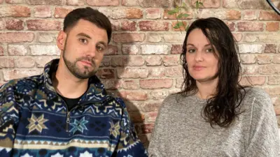 A dark-haired man, wearing a colourful fleece, and a dark-haired woman, wearing a grey top, stand against an indoor brick wall