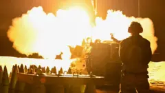 An Israeli soldier in Gaza, surrounded by artillery shells, watches a missile being launched.