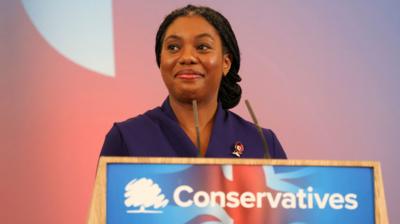 Kemi Badenoch in a purple suit standing behind a podium with the Conservative party logo