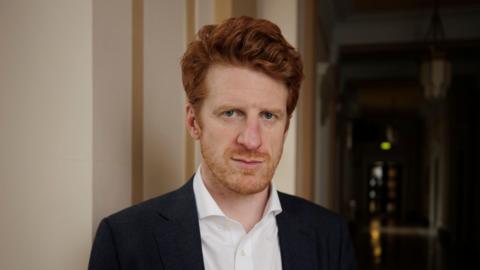 Matthew O'Toole wearing a black suit jacket and a white shirt. He has ginger hair and a ginger beard.