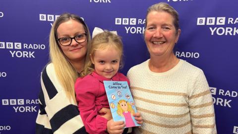 Willow, pictured with her mum Rachel and author Sylvia Buck, holds a copy of a book entitled Willow Come to Tea? against a purple background.