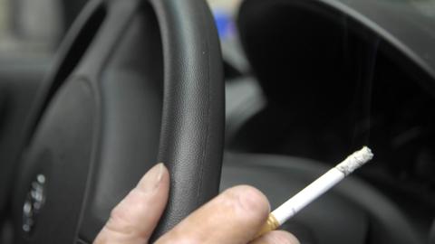 Close up of a hand holding a steering wheel and lit cigarette.