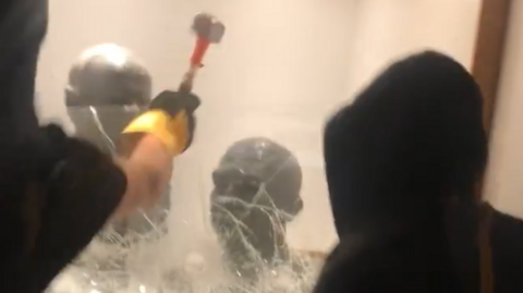 screengrab of two dark-hooded people using a hammer to break a display case with two busts of Chaim Weizmann