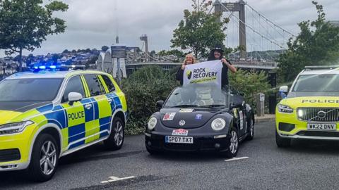 A man and woman stand on the front seats of an open top Volkswagen Beetle, holding a sign saying Rock 2 Recovery. The car is flanked by two police cars and the Tamar bridge is in the background.