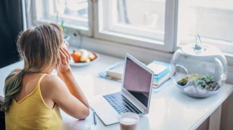 Young woman gazing out window sitting at desk in front of laptop, with fruit, books and plants in a glass sphere in front of her