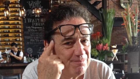 A man looking straight at the camera, with his index finger resting on his temple and a pair of glasses across his forehead. He is sitting in a restaurant. A barman is standing behind a bar in the background, making a cocktail.