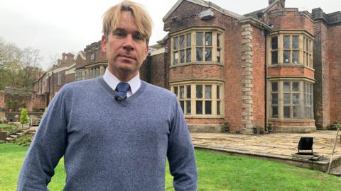 Hopwood DePree stands outside Hopwood Hall, in Middleton, Rochdale. He has blond hair and his wearing a tie under a blue jumper. 