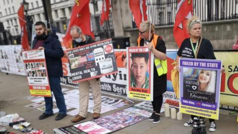 Families in London protesting at the number of mental health deaths in Essex. There are two men and two women holding placards with pictures of their loved ones, alongside words including "failed by the state".