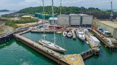 A photo which shows some of Pendennis Shipyard from an aerial view. There is sea and water in the distance which is a bluey-green colour. There is also a helicopter landing pad with a yellow circle around the letter H. 