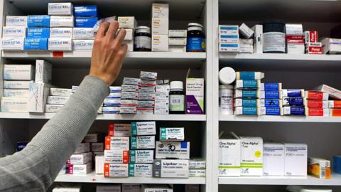 A person with a grey jumper reaches for medicine in a pharmacy shelf. There are a number of different medicines boxes and bottles stacked in the shelves.