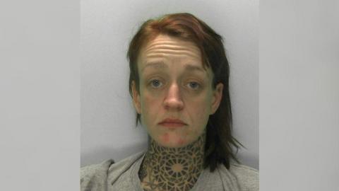 Mugshot of Hannah Roberts - a woman in her early 30s with a distinctive, tribal-style tattoo covering her neck. She has medium length dyed red hair, and is wearing a grey tracksuit.